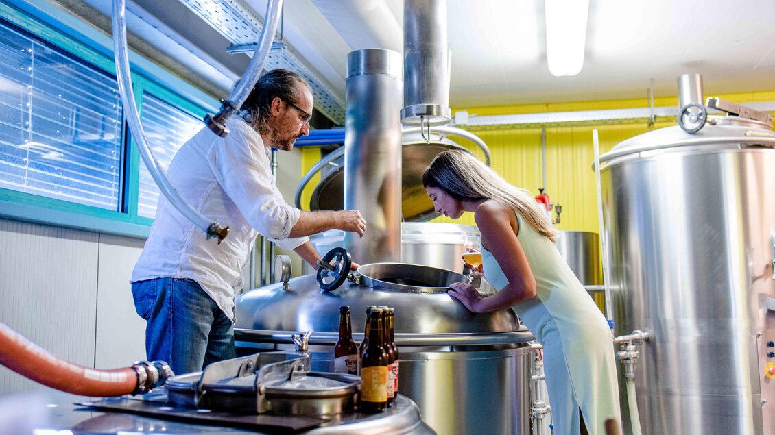 Ein Mann und eine Frau stehen in einer Brauerei neben großen, silbernen Braukesseln. Sie beobachten den Brauprozess und scheinen interessiert an der Herstellung von Bier zu sein. | © Lava Bräu