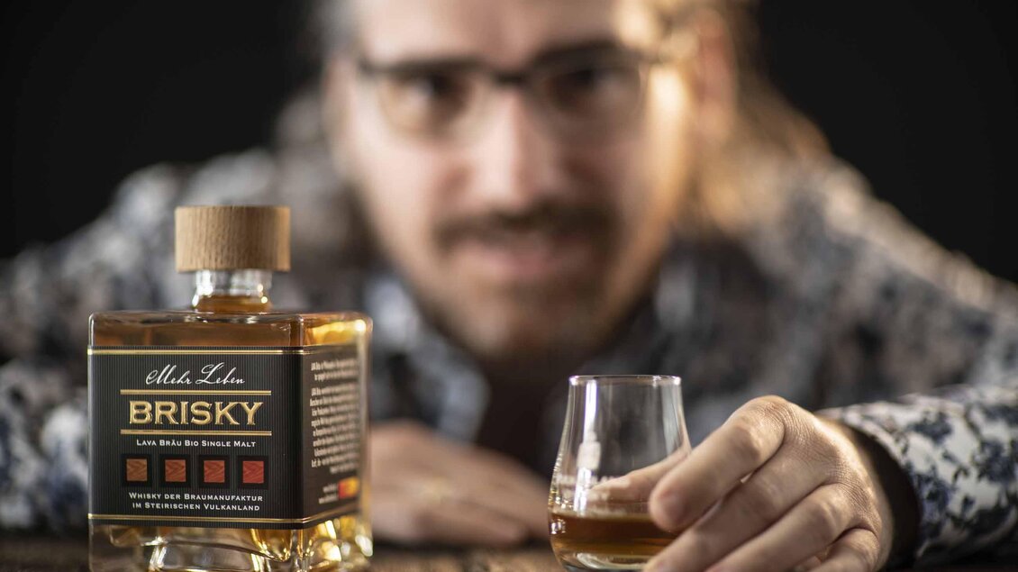 Ein Mann mit Brille hält ein Glas Whisky in der Hand. Im Vordergrund steht die Flasche Brisky Whisky auf einem Holztisch. | © Lava Bräu