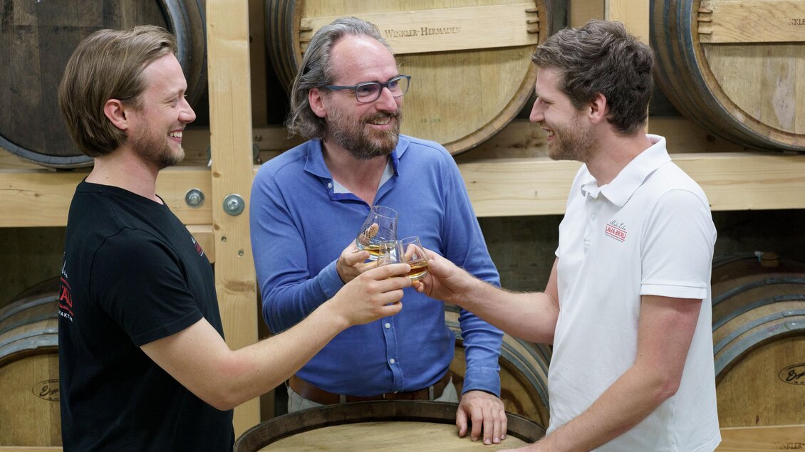 Drei Männer stoßen mit Gläsern an und feiern in einer Umgebung mit Holzfässern. Die Stimmung ist freundlich und gesellig.