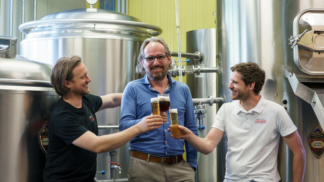 Drei Männer stoßen mit Biergläsern in einer Brauerei an. Im Hintergrund sind große, silberne Brautanks zu sehen.