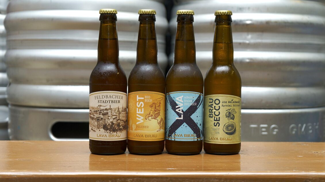 Vier Flaschen Bier mit unterschiedlichen Etiketten stehen auf einem Tisch. Im Hintergrund sind Metallfässer zu sehen.