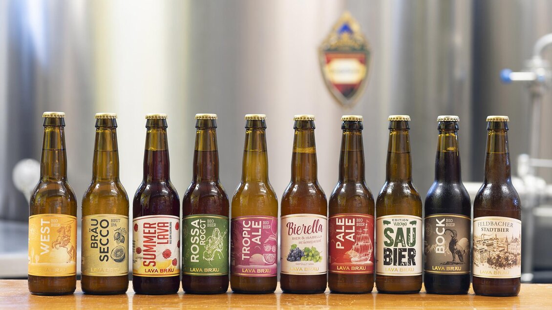Eine Reihe von Bierflaschen mit verschiedenen Etiketten steht auf einer Holztheke. Im Hintergrund sind silberne Brauausrüstungen sichtbar.