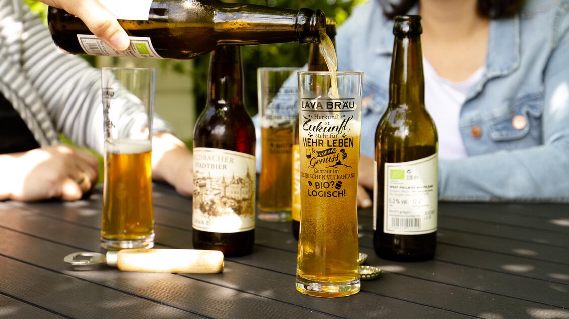 Ein Glas Bier wird ausgegossen, während weitere Bierflaschen auf einem Tisch stehen. Im Hintergrund sind Menschen zu sehen, die entspannt sitzen.