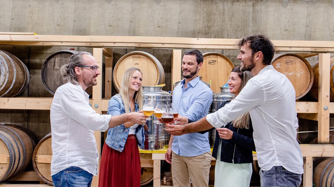 Eine Gruppe von fünf Personen stößt mit Gläsern an. Im Hintergrund sind Holzfässer zu sehen.