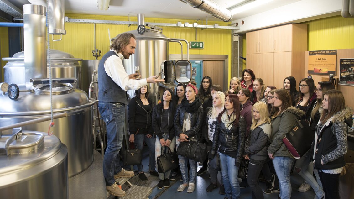 Eine Gruppe von Frauen hört einem Mann zu, der ihnen in einer Brauerei etwas erklärt. Im Hintergrund sind große Metallbehälter und eine moderne Einrichtung zu sehen.