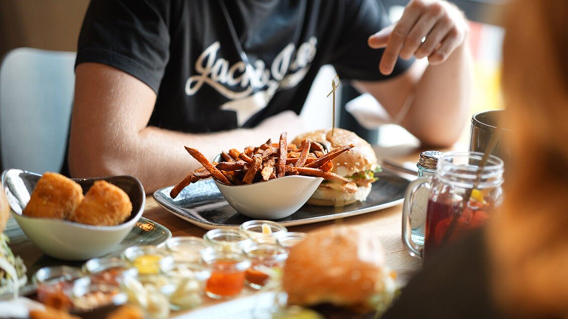 Ein Tisch mit köstlichem Essen, einschließlich eines Burgers und süßen Kartoffel-Pommes. Im Hintergrund sind verschiedene Beilagen und Getränke zu sehen. | © Le Burger - The Guardians
