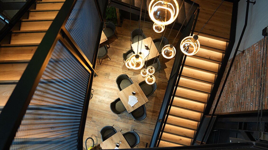 Eine moderne Treppe mit eleganten Leuchten führt zu einem gemütlichen Essbereich. Der Raum ist hell und einladend, mit Holz- und Stahlelementen. | © Le Burger - The Guardians