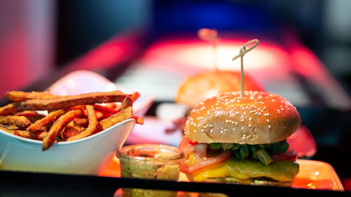 Ein saftiger Burger mit Sesambrötchen und knusprigen Süßkartoffel-Pommes. Im Hintergrund ist eine moderne Restaurantatmosphäre mit sanfter Beleuchtung zu sehen. | © Le Burger - The Guardians