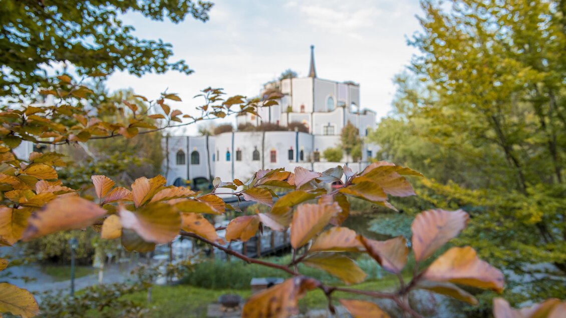 Ein schöner Blick auf ein Gebäude umgeben von herbstlichen Bäumen und bunten Blättern. Die ruhige Atmosphäre wird durch das Wasser im Vordergrund verstärkt. | © Kurkommission Bad Blumau