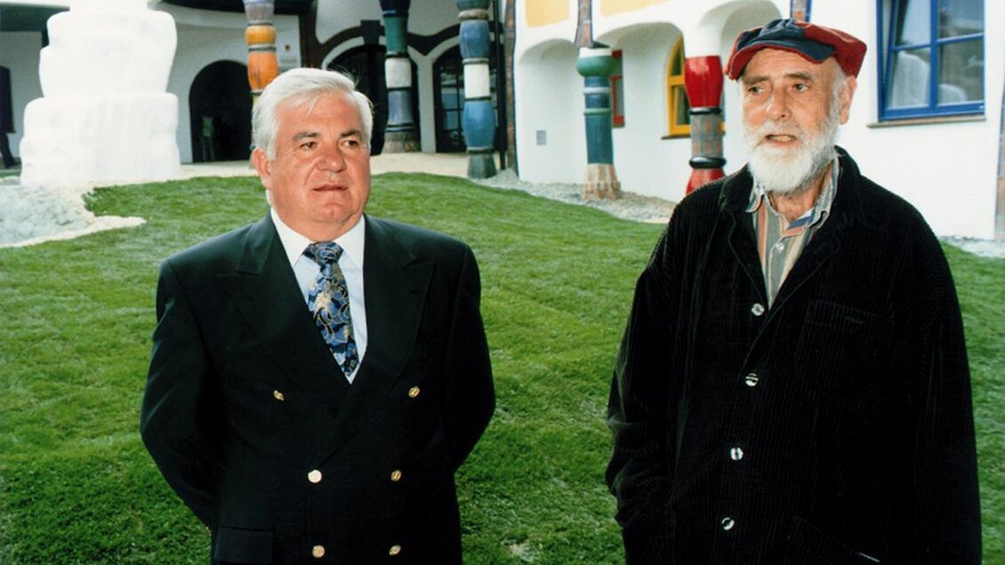 Zwei Männer stehen in einem gepflegten Garten vor einem farbenfrohen Gebäude. Der Mann links trägt einen Anzug, während der Mann rechts einen Kapuzenpulli und eine Mütze trägt. | © Rogner Bad Blumau