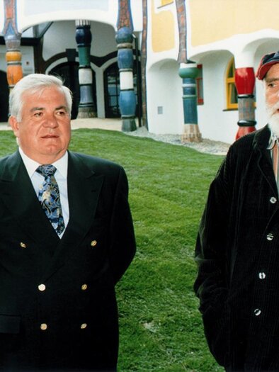 Zwei Männer stehen in einem gepflegten Garten vor einem farbenfrohen Gebäude. Der Mann links trägt einen Anzug, während der Mann rechts einen Kapuzenpulli und eine Mütze trägt. | © Rogner Bad Blumau