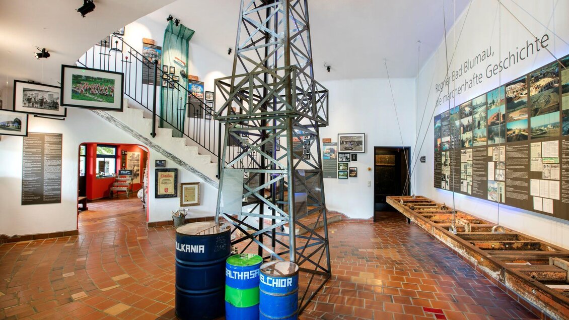 Ein Innenraum eines Museums mit einer Treppe und Ausstellungsplatten an der Wand. In der Mitte steht eine Nachbildung einer Ölbohrinsel mit zwei Fässern. | © Rogner Bad Blumau
