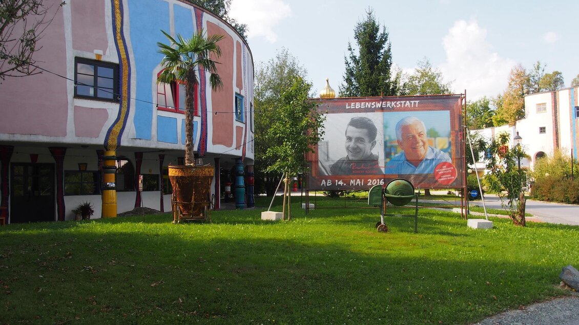 Ein buntes Gebäude mit Pflanzen und grüner Wiese. Ein großes Plakat mit zwei Porträts steht im Vordergrund. | © Rogner Bad Blumau