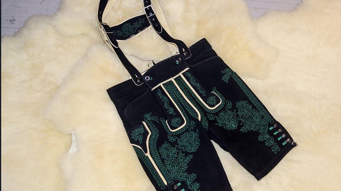 Eine traditionelle bayerische Lederhose mit grüner Stickerei und schwarzen Trägern. Sie liegt auf einem hellen, pelzigen Untergrund. | © Leder Traninger