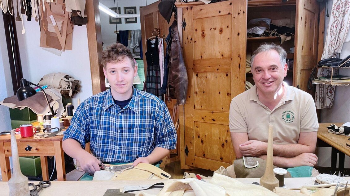 Zwei Männer arbeiten in einer Werkstatt. Sie sitzen an einem Tisch mit Materialien und Werkzeugen zur Lederbearbeitung. | © Christian Raich