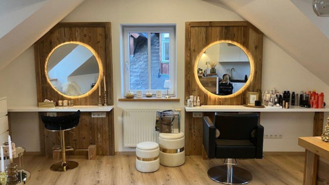 Ein moderner Friseursalon mit großen, beleuchteten Spiegeln und stylischen Stühlen. Die Wände sind aus Holz, und es gibt verschiedene Pflegeprodukte auf den Regalen. | © TVB Ausseerland- Salzkammergut_Heike Temmel