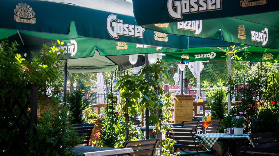 Ein gemütlicher Biergarten mit grünen Sonnenschirmen und vielen Pflanzen. Die Tische sind ordentlich gedeckt und laden zum Verweilen ein.