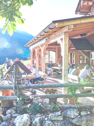 Eine einladende Terrasse mit Holzverkleidung, auf der Menschen in der Sonne sitzen und zwischen den Bergen entspannen. Die Umgebung ist grün und freundlich, ideal für gesellige Zusammenkünfte. | © Lenzbauer Alm