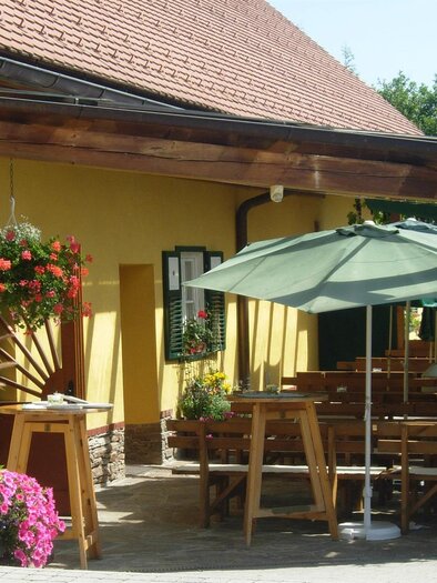 Ein einladendes Gebäude mit gelber Fassade und einem großen Außensitzbereich. Es gibt blühende Pflanzen und einen Sonnenschirm, der Schatten spendet. | © Fam. Peter