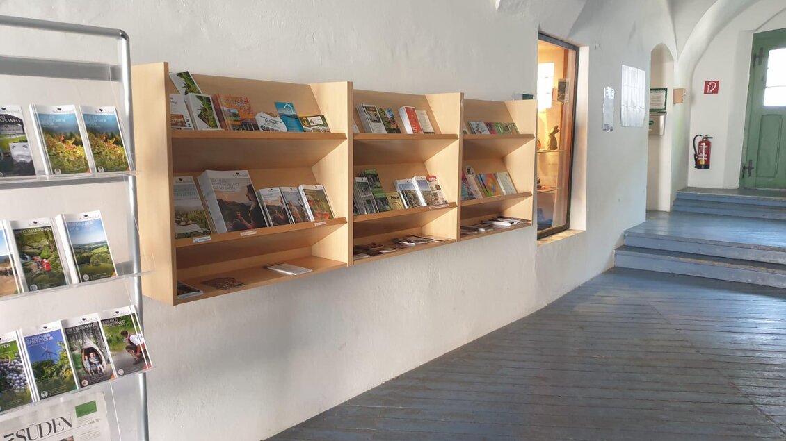 Ein heller Flur mit Holzregalen, die mit verschiedenen Broschüren gefüllt sind. Auf der linken Seite steht ein Ständer mit weiteren Informationsmaterialien. | © Sabrina Prattes
