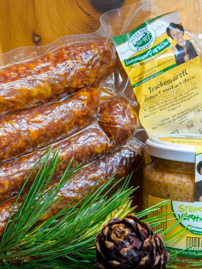 A package of sausage and a jar of mustard are placed on a wooden surface. Next to the food, there are some fir branches and a pine cone. | © Erlebnisregion Murtal