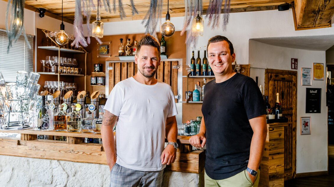 Zwei Männer stehen an der Bar eines gemütlichen Lokals. Im Hintergrund sind Flaschen und dekorative Elemente sichtbar. | © Region Graz - Mias Photoart