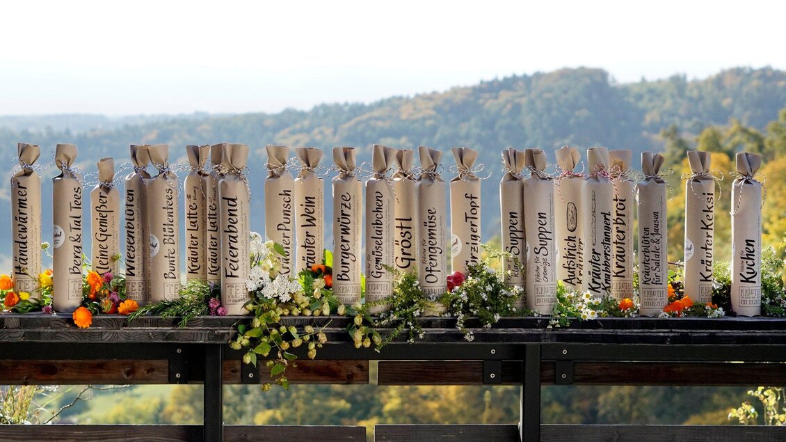 Eine Reihe von dekorierten Flaschen steht auf einem Tisch, umgeben von Blumen und grünem Laub. Im Hintergrund sieht man sanfte Hügel und einen klaren Himmel. | © Sabine Lienhart