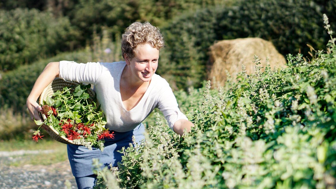 Eine Frau erntet frische Kräuter in einem Garten. Im Hintergrund sind Heuballen und grüne Pflanzen zu sehen. | © Sabine Lienhart