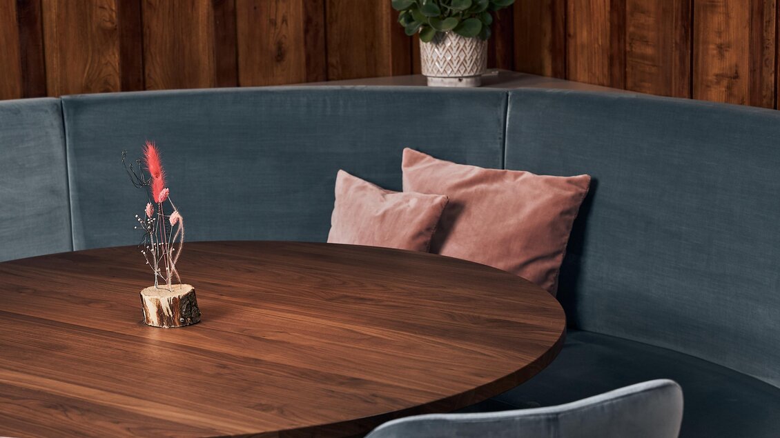 Ein stilvoller Tisch aus Holz mit einer modernen, runden Bank und dekorativen Kissen. Eine grüne Pflanze fügt einen frischen Akzent hinzu.