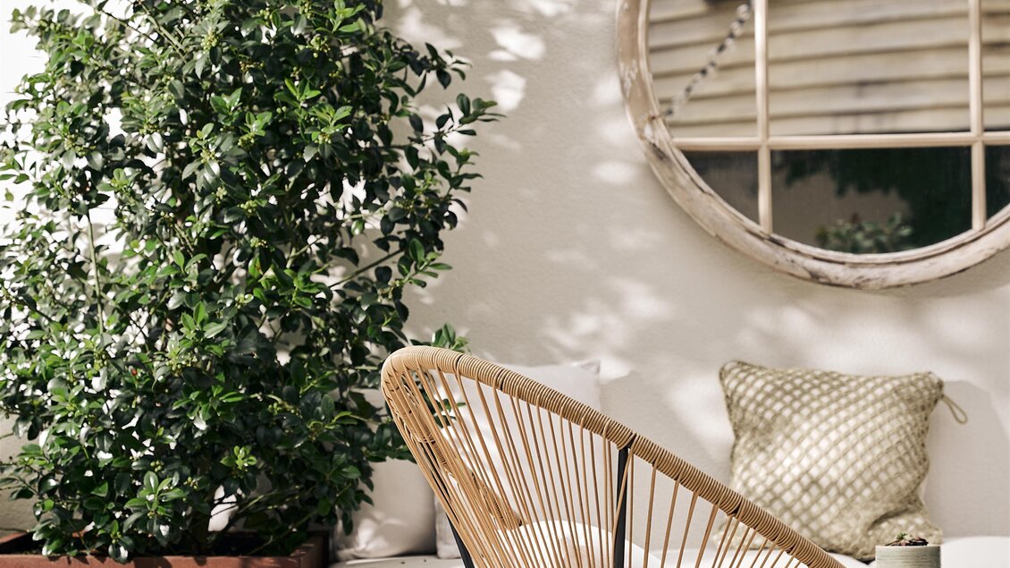 Ein stilvoller Außenbereich mit einem Korbsessel und einer großen Pflanze in einem Topf. Im Hintergrund befindet sich ein runder Spiegel und eine Holzoberfläche.