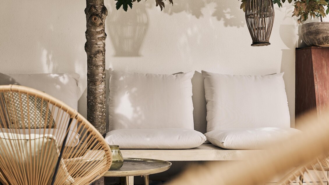 Ein eleganter Wohnbereich mit bequemen Kissen und einem Tisch. Im Hintergrund sind Pflanzen und hängende Lampen zu sehen.