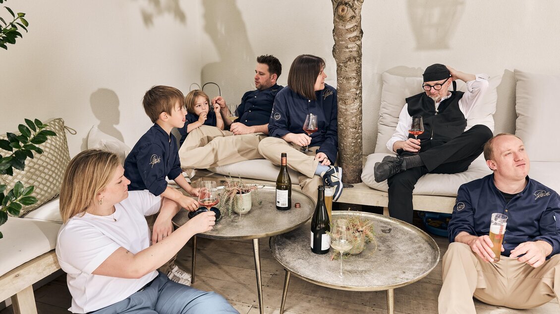 Eine Gruppe von Menschen sitzt entspannt auf einem Sofa in einem modernen Raum. Sie genießen Getränke und unterhalten sich miteinander.