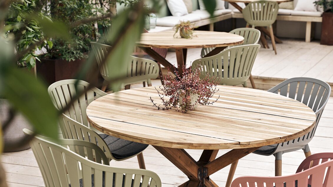 Ein gemütlicher Außenbereich mit einem runden Holztisch und bunten Stühlen. Pflanzen dekorieren die Umgebung und schaffen eine einladende Atmosphäre.