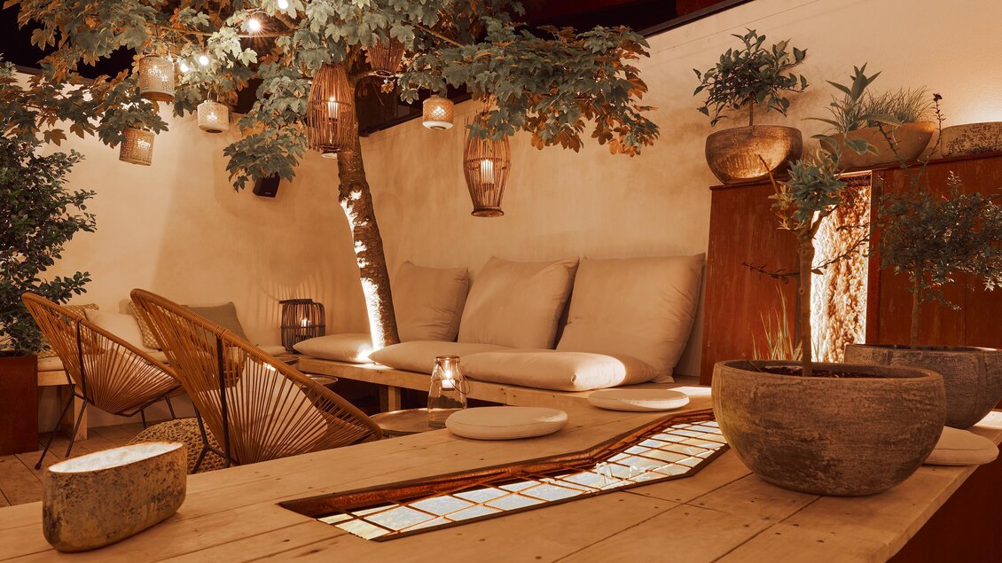 Ein gemütlicher Raum mit Holzdetails, Pflanzen und weichen Sitzgelegenheiten. Die warmen Lichter schaffen eine einladende Atmosphäre.