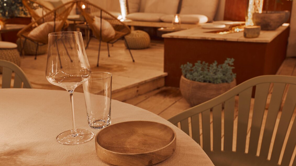 Ein eleganter Tisch mit einem Weinglas, einem Wasserglas und einer Holzschale. Im Hintergrund sind gemütliche Sitzgelegenheiten und sanfte Beleuchtung zu sehen.