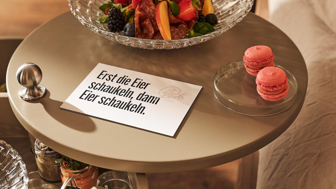 Ein Tisch mit einer Schüssel frischem Obst und zwei Rosa-Macarons. Neben dem Obst liegt ein Blatt Papier mit dem Spruch: "Erst die Eier schaukeln, dann Eier schaukeln."