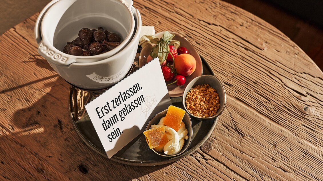 Eine Auswahl an Snacks auf einem Holztablett, darunter Früchte und Nüsse. Ein Schild mit der Aufschrift „Erst zulassen, dann gelassen sein“ liegt daneben.