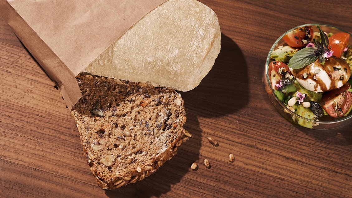 Ein frisches Vollkornbrot liegt auf einem Holztisch, teilweise aus der Verpackung genommen. Daneben befindet sich eine Schüssel mit einem bunten Salat.