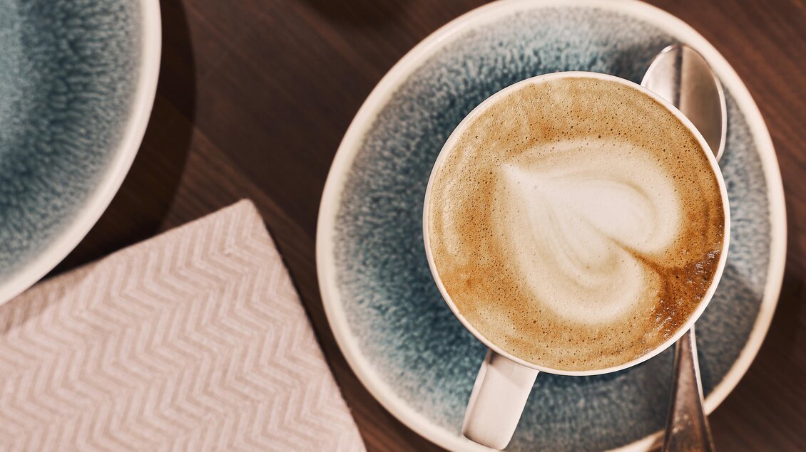 Eine Tasse Cappuccino mit einem Herz-Motiv aus Milchschaum auf einem blauen Unterteller. Daneben liegt ein kleiner Löffel und eine Serviette.