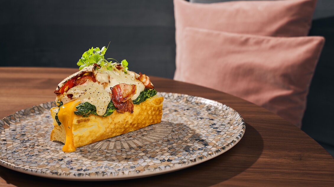 Ein köstliches Gericht auf einem dekorativen Teller, das mit frischem Gemüse und Käse belegt ist. Der Hintergrund zeigt eine stilvolle Einrichtung mit weichen Kissen.