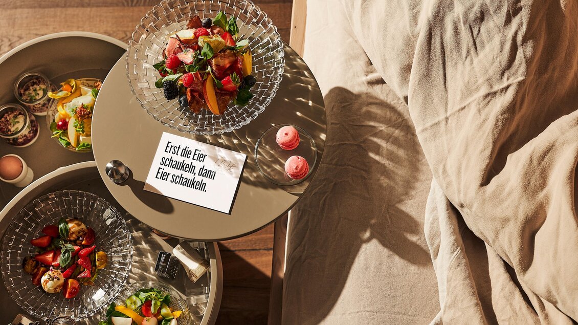 Ein stilvoller Tisch mit verschiedenen gesunden Speisen und einem Zettel. Im Hintergrund sieht man ein Bett mit einer gemütlichen Bettdecke.