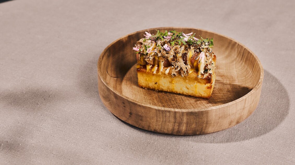 Ein Stück Brot mit zartem Fleisch und frischen Kräutern, serviert auf einem Holzbrett. Die Präsentation ist schlicht und ansprechend.
