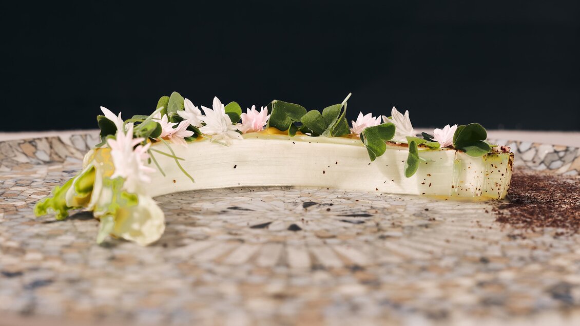 Ein kunstvoll angerichteter Teller mit einer eleganten Speise, dekoriert mit frischen Kräutern und essbaren Blüten. Die Präsentation ist modern und ansprechend, auf einem stilvollen Geschirr.