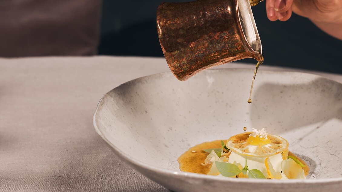 Eine elegant angerichtete Speise auf einem Teller. Jemand gießt eine goldene Soße darüber.