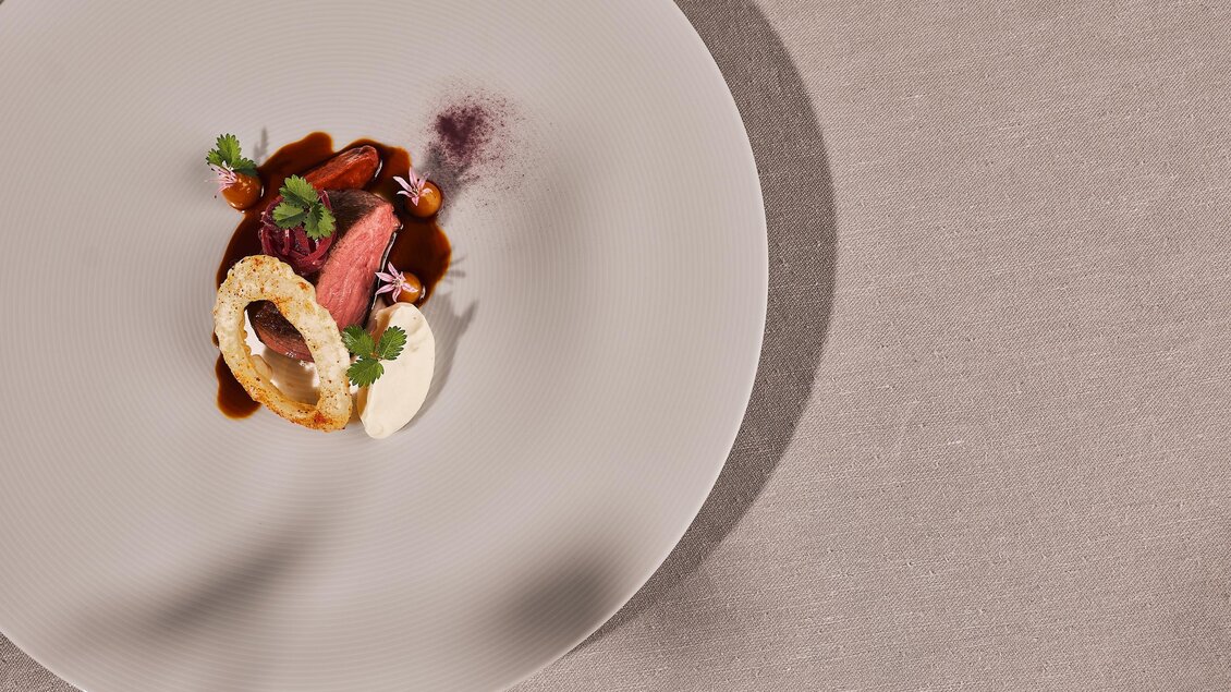 Ein kunstvoll angerichtetes Gericht auf einem weißen Teller. Es enthält Fleisch, eine Sauce und frische Kräuter.