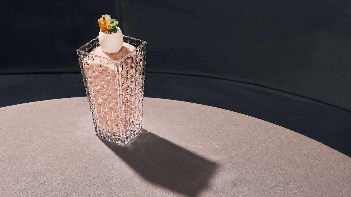 Ein eleganter Cocktail in einem Kristallglas steht auf einem runden Tisch. Der Drink ist mit einer dekorativen Frucht und einem Minzblatt verziert.