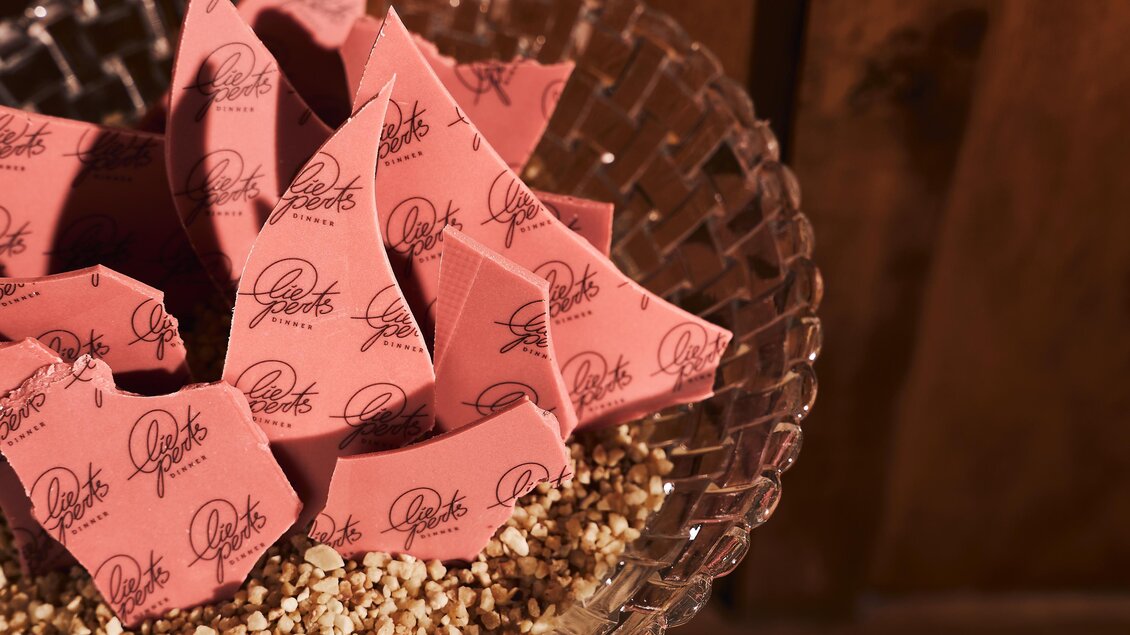 Ein Glasbehälter mit zerbrochenen rosa Schokoladenteilen, die auf einer Schicht aus Nüssen liegen. Die Schokolade ist mit einem Logo bedruckt.