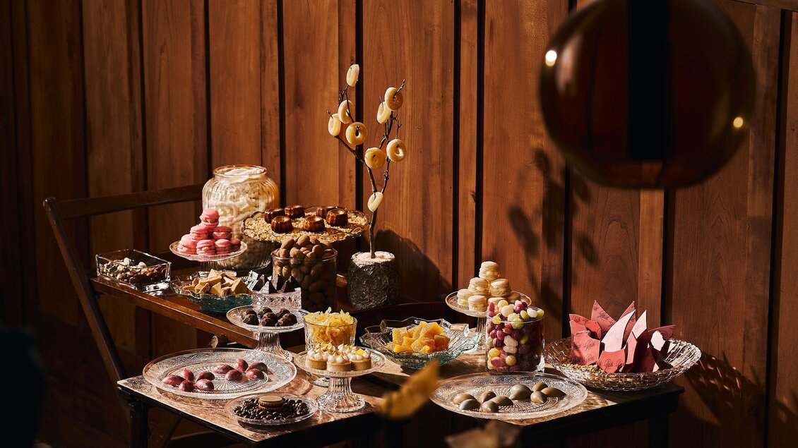 Ein Tisch mit einer Vielzahl von Süßigkeiten und Desserts ist dekoriert. Im Hintergrund sind elegante Holzpaneele zu sehen.