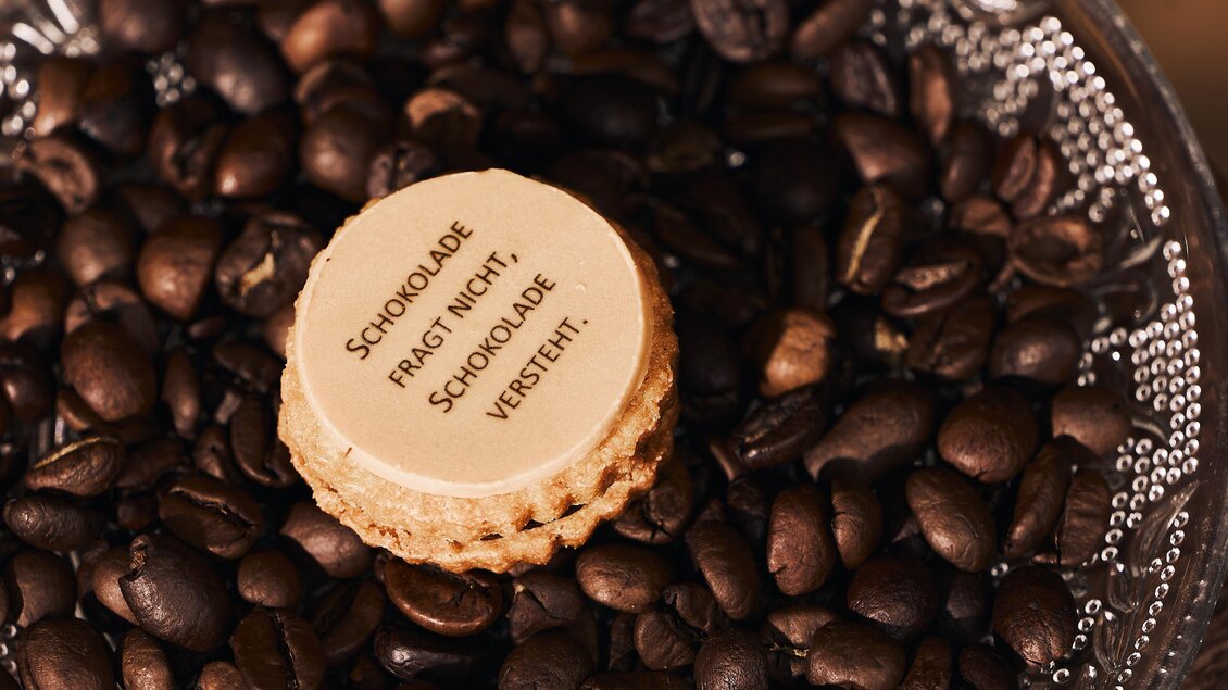 Eine Schokoladenverpackung liegt auf einer Schicht von gerösteten Kaffeebohnen. Der Text auf der Verpackung lautet: "Schokolade fragt nicht, Schokolade versteht."