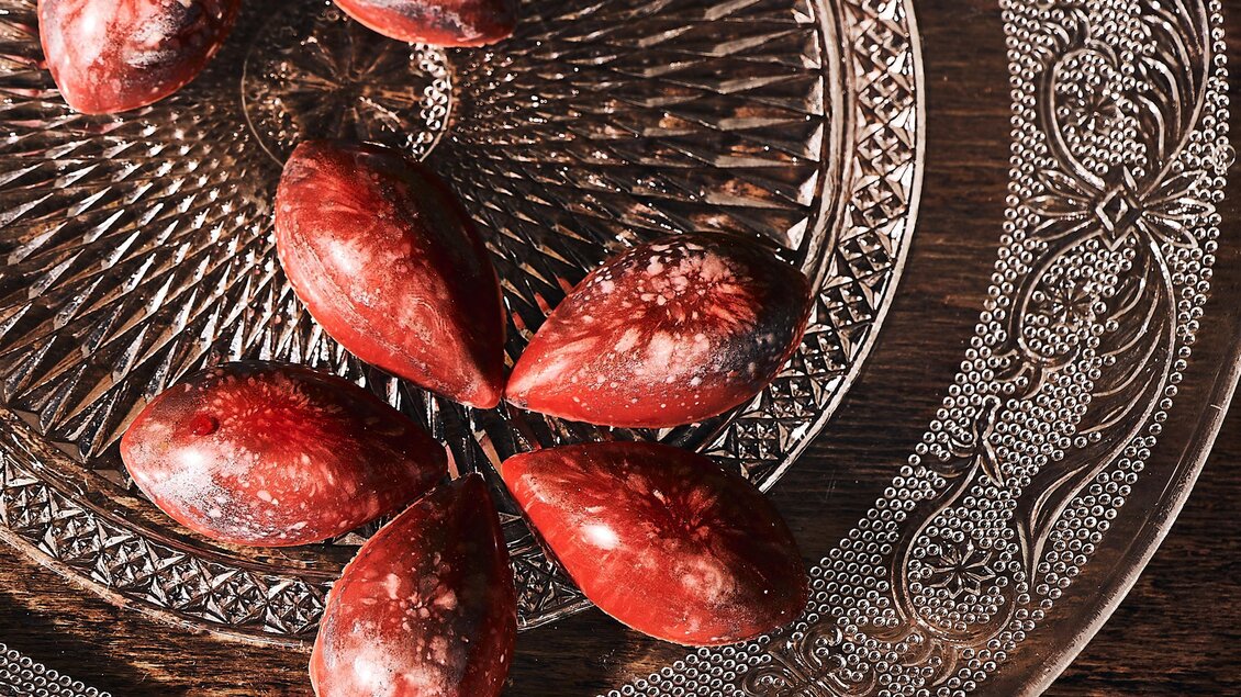 Eine dekorative Glasschale mit einer Anordnung von roten, tropfenförmigen Objekten. Die Schale hat ein filigranes Muster und ist auf einem Holzuntergrund platziert.